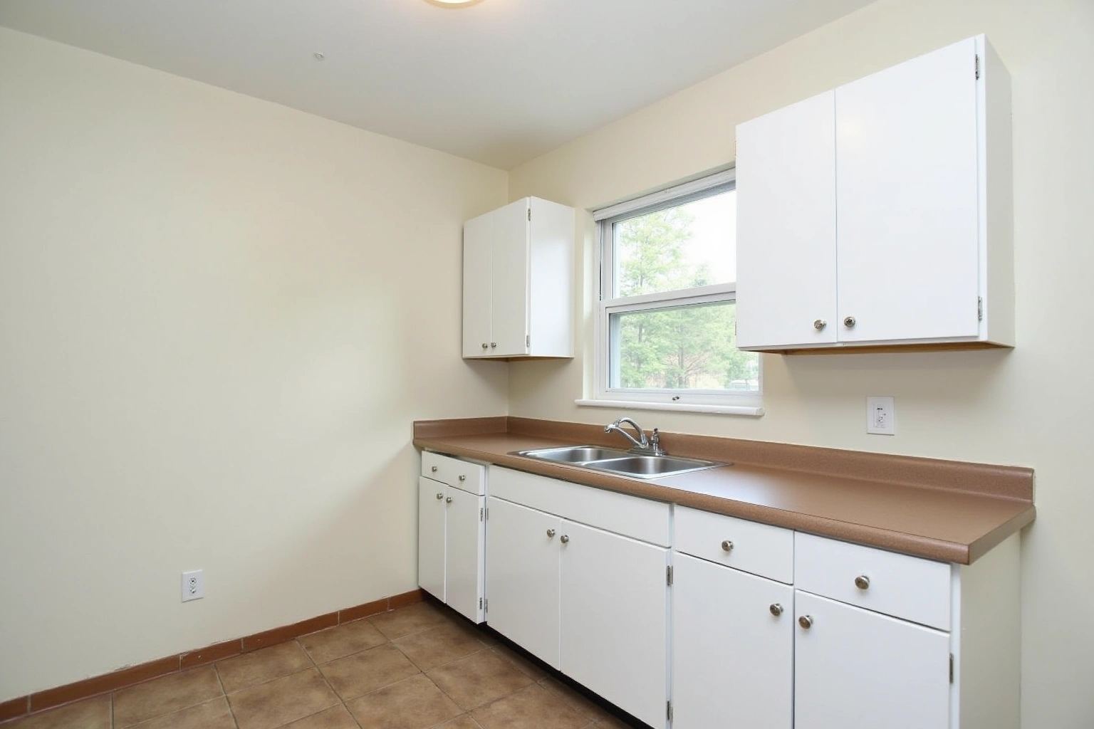 Plain white flat-panel cabinets with a brown laminate counter, a stainless sink under a window and tan tile floor.