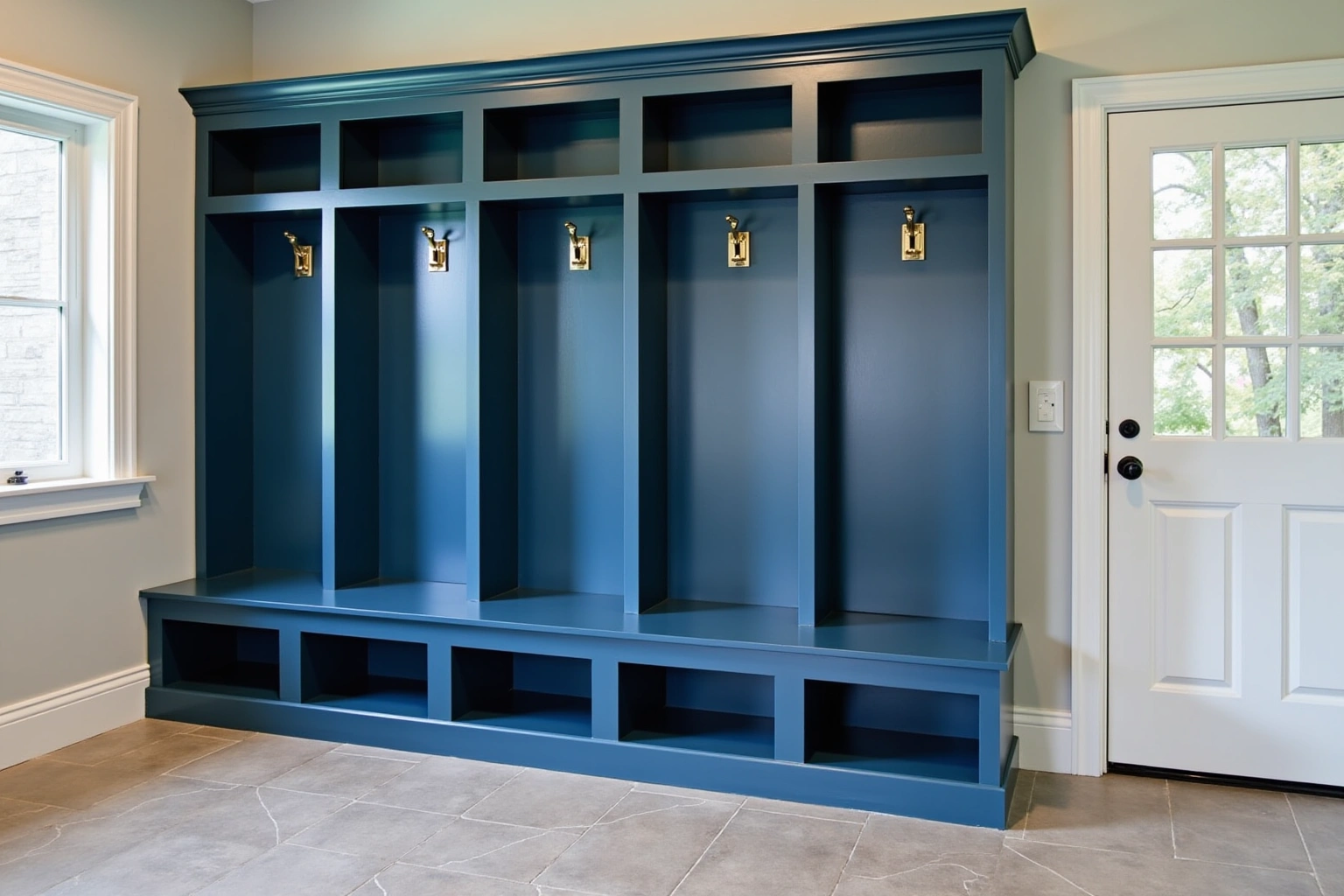 Five deep blue mudroom lockers with brass hooks and open cubbies above a long bench, next to a glazed door.
